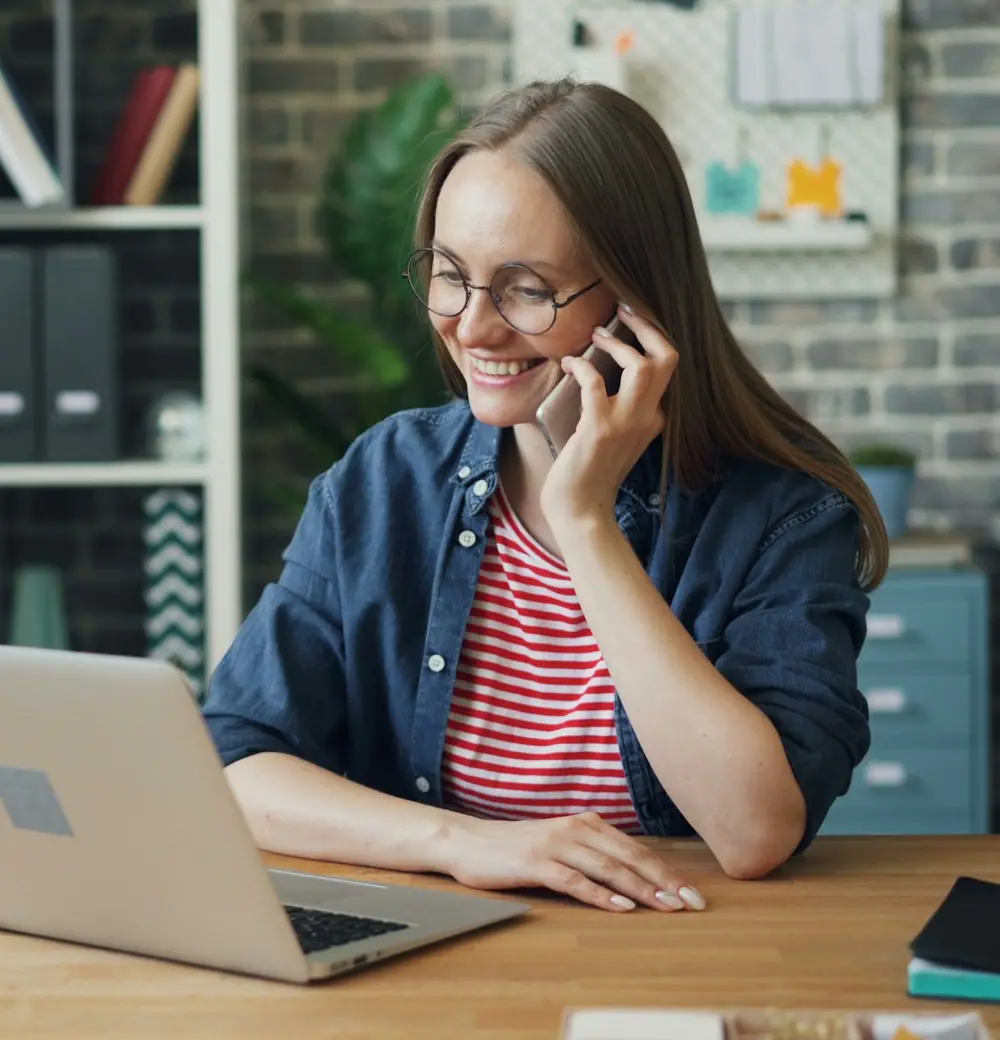 A woman in glasses laughing on the phone at her desk, laptop open