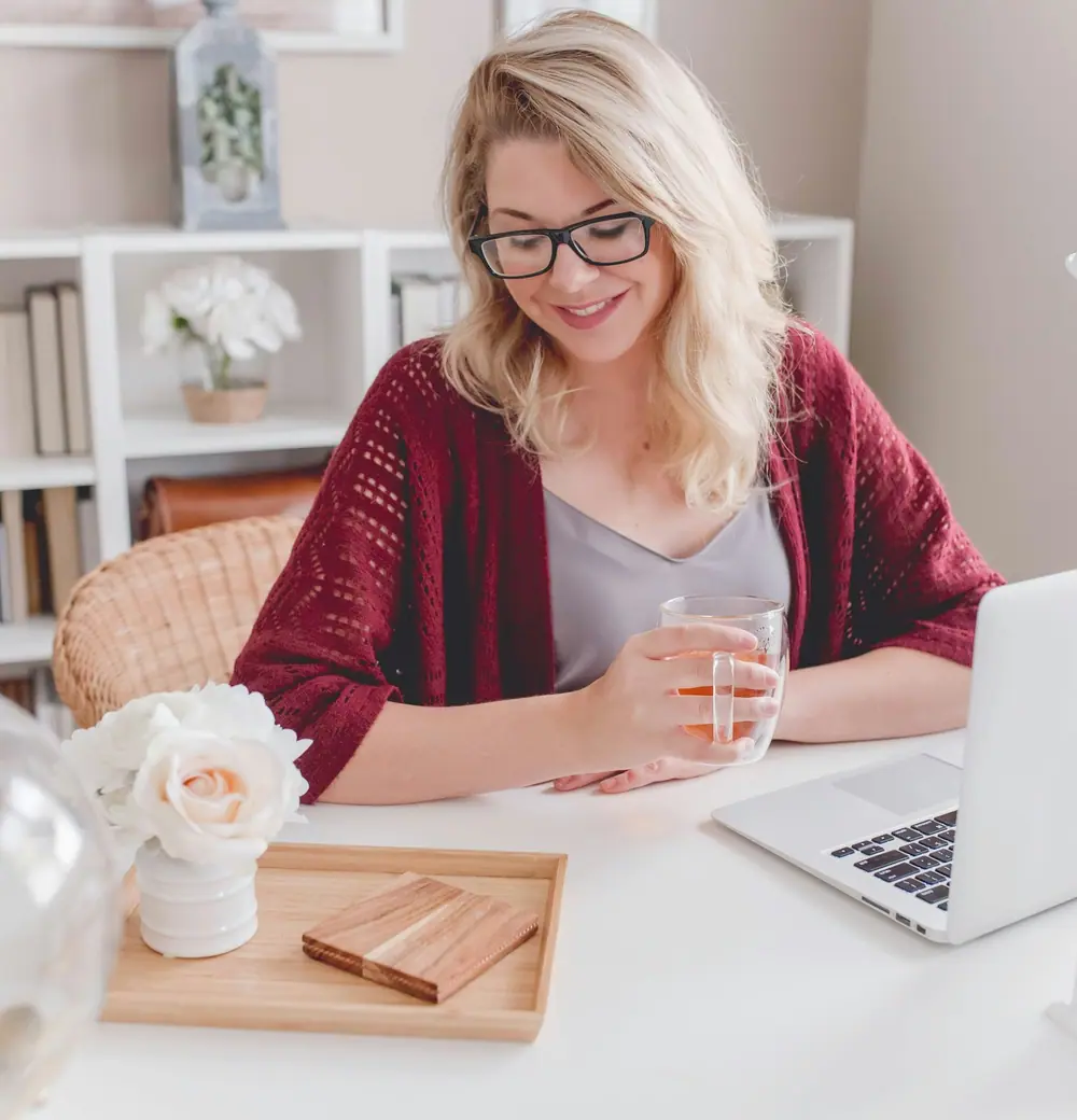 A woman smiling at her laptop with a cup in her hand