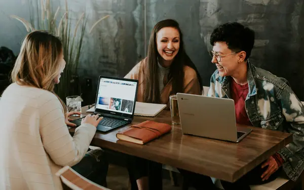 Three colleagues laughing together around laptops at a wooden table