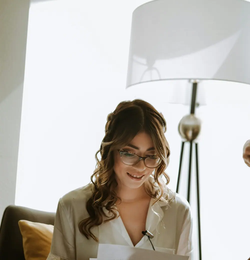 A woman in glasses smiling as she reads at a bright desk
